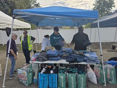 Volunteers handing out folded jeans and clothing from a table under a canopy, with bags of clothes lined up underneath